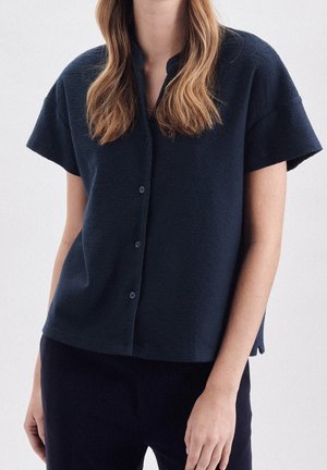 Kobieta ubrana w granatową teksturowaną koszulę na guziki z krótkim rękawem o luźnym kroju oraz ciemne spodnie, pokazana od szyi do bioder.