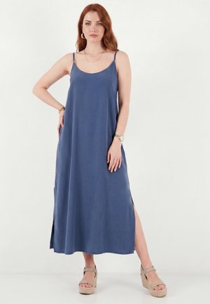 Frau mit roten Haaren, die ein ärmelloses blaues Midikleid mit seitlichen Schlitzen und beigen Keilsandalen trägt, steht vor einem weißen Hintergrund.
