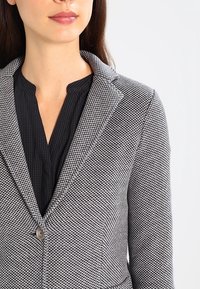 Blazer a scacchi grigi e neri con rever a punta, chiusura con un solo bottone e tessuto testurizzato. Indossato sopra una blusa scura e plissettata.