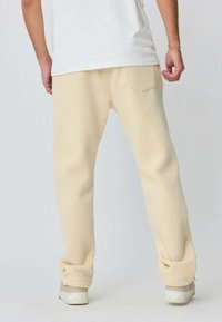 Pantalon de survêtement beige avec une coupe décontractée, des poignets élastiques et deux poches arrière, associé à des baskets de couleur claire, vu de l'arrière.