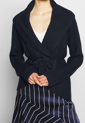 Femme portant un pull en maille noir côtelé, portefeuille, avec un large col et une ceinture, associé à une jupe rayée bleu marine et blanche.