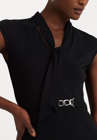 Černé šaty se zavazovacím výstřihem, krátkými rukávy a detaily stříbrného pásu z řetízkových oček. Hladká látka s mírným leskem.