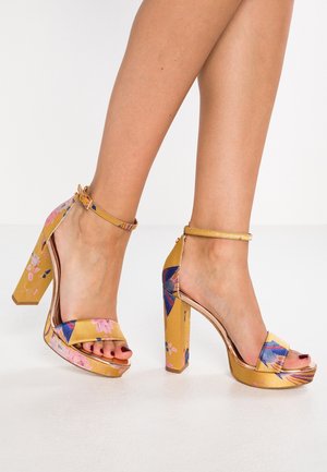 Sandały na obcasie z kwiatowym wzorem w kolorze żółtym, z grubym obcasem, paskiem na kostkę i krzyżującym się przodem. Teksturowana tkanina.