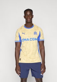 Μπλούζα ποδοσφαίρου Puma σε μπεζ χρώμα με μπλε λεπτομέρειες, που έχει σχέδιο αστικού τοπίου και το λογότυπο του συλλόγου. Φοριέται με σκούρα μπλε σορτς, παρουσιάζοντας ένα αθλητικό σχέδιο.