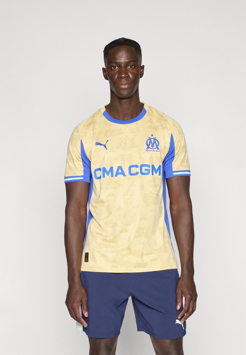 Μπλούζα ποδοσφαίρου Puma σε μπεζ χρώμα με μπλε λεπτομέρειες, που έχει σχέδιο αστικού τοπίου και το λογότυπο του συλλόγου. Φοριέται με σκούρα μπλε σορτς, παρουσιάζοντας ένα αθλητικό σχέδιο.