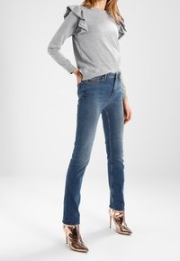 Harmaa collegepaita, jossa on röyhelöyksityiskohtia hihoissa, yhdistetty tiukkoihin sinisiin farkkuihin ja kiiltäviin ruusukultaisiin korkeakorkoisiin saappaisiin.