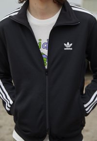 Svart zip-up träningsjacka med vitt Adidas-logga och tre vita ränder på varje ärm. Bärs över en vit grafisk t-shirt. Slät textur.