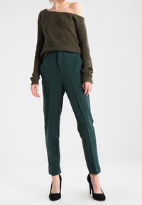Ciemnozielone dopasowane spodnie zestawione z oliwkowym swetrem bez ramiączek z prążkowanej tkaniny. Modelka nosi czarne szpilki, a sweter ma długie rękawy.