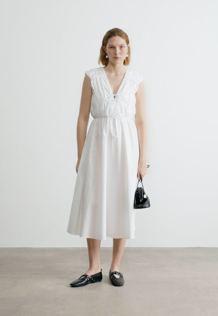 Femme portant une robe midi blanche sans manches, des ballerines noires, tenant un petit sac à main noir, debout contre un mur blanc uni.