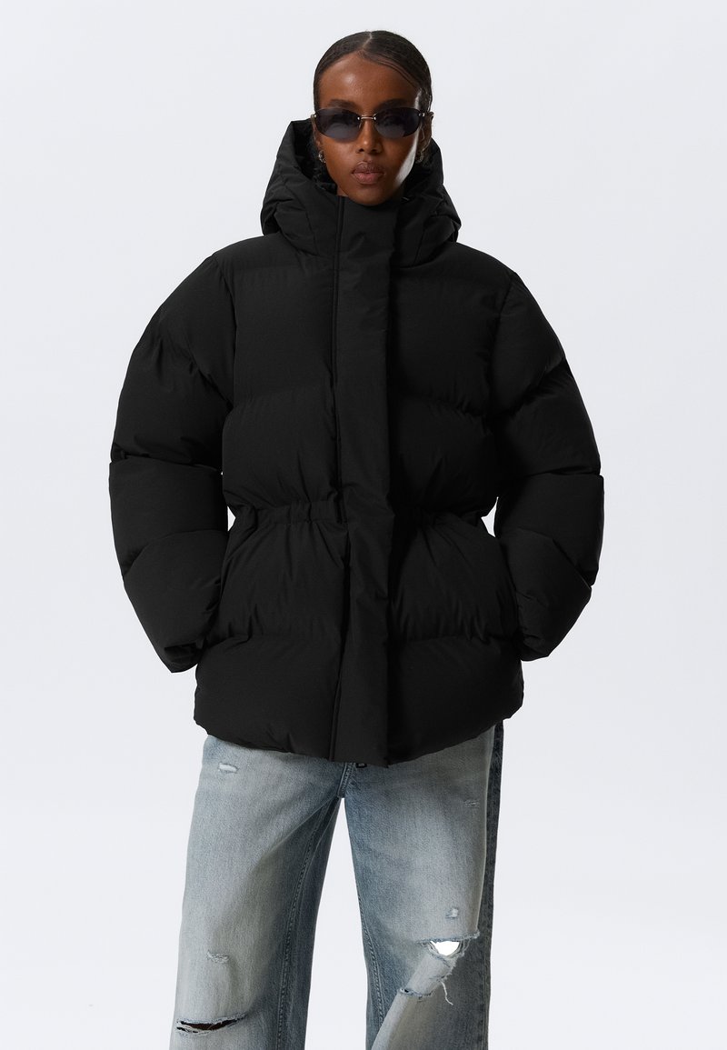Schwarze Pufferjacke mit hohem Kragen, elastischem Taillenbund und übergroßer Silhouette. Kombiniert mit ausgewaschenen hellblauen Jeans.