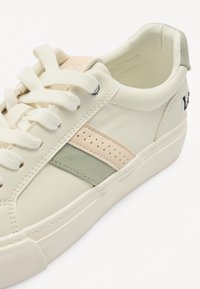 Zapatilla de cuero color crema con una raya de acento en verde suave y durazno claro, superficie texturizada y cordones planos color beige. Detalles de diseño perforado.