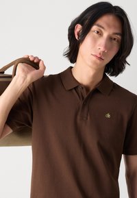 Braunes Poloshirt mit kurzen Ärmeln, zwei Knöpfen und einem grünen gestickten Logo. Das Modell hält eine beige Tasche über einer Schulter.
