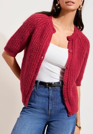 Adīts sarkans kardigans ar īsām piedurknēm, ribota tekstūra un pieci pogas priekšpusē. Valkāts virs baltas blūzes un kombinēts ar augsta vidukļa zilām džinsām.