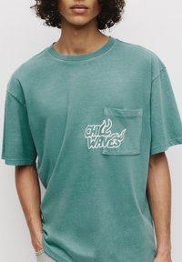 Teal kortærmet t-shirt lavet af blødt stof, med en brystlomme og hvidt grafisk tekst "CHILL WAVES" med flamme accenter.