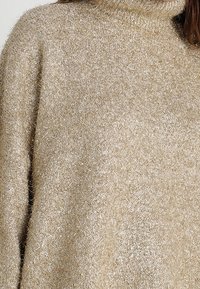 Beżowy sweter z golfem wykonany z miękkiej, teksturowanej tkaniny z delikatnymi metalicznymi włóknami. Posiada luźny krój i obszerne rękawy.