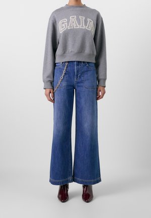 Person trägt ein graues kurzes Sweatshirt mit dem Schriftzug „GAIA“, weite blaue Jeans mit einem Kettenaccessoire und bordeauxrote spitze Stiefel.