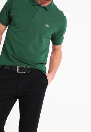 Homme portant un polo vert avec un petit logo crocodile, pantalon noir et ceinture noire, main dans la poche.