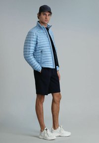 Veste matelassée bleu clair avec un motif de piquage vertical, poches zippées, associée à un short bleu marine et des baskets blanches. Modèle se tenant de côté.
