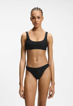 Frau mit geflochtenem Haar, die einen schwarzen, strukturierten Bikini mit einem Oberteil mit U-Boot-Ausschnitt und hochgeschnittenen Höschen trägt, steht vor einem weißen Hintergrund.