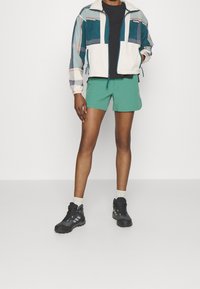 Kolorowa kurtka z geometrycznym wzorem, czarna koszulka, zielone szorty i czarne buty trekkingowe z szarymi akcentami. Spójny strój na zewnątrz.