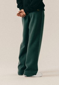 Osoba w ciemnozielonych szerokich spodniach i białych sneakersach stoi z rękami złożonymi za plecami na neutralnym tle.