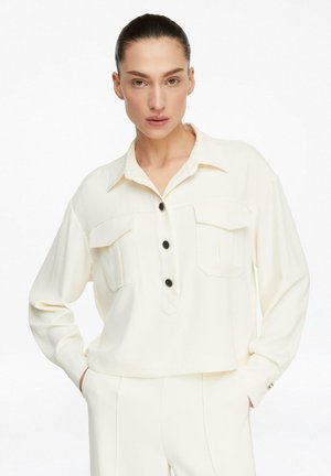 Frau mit nach hinten gegelten Haaren, die ein elfenbeinfarbenes Hemd mit Taschen und passenden, hoch taillierten Hosen trägt, Hände in den Taschen, neutraler Ausdruck.