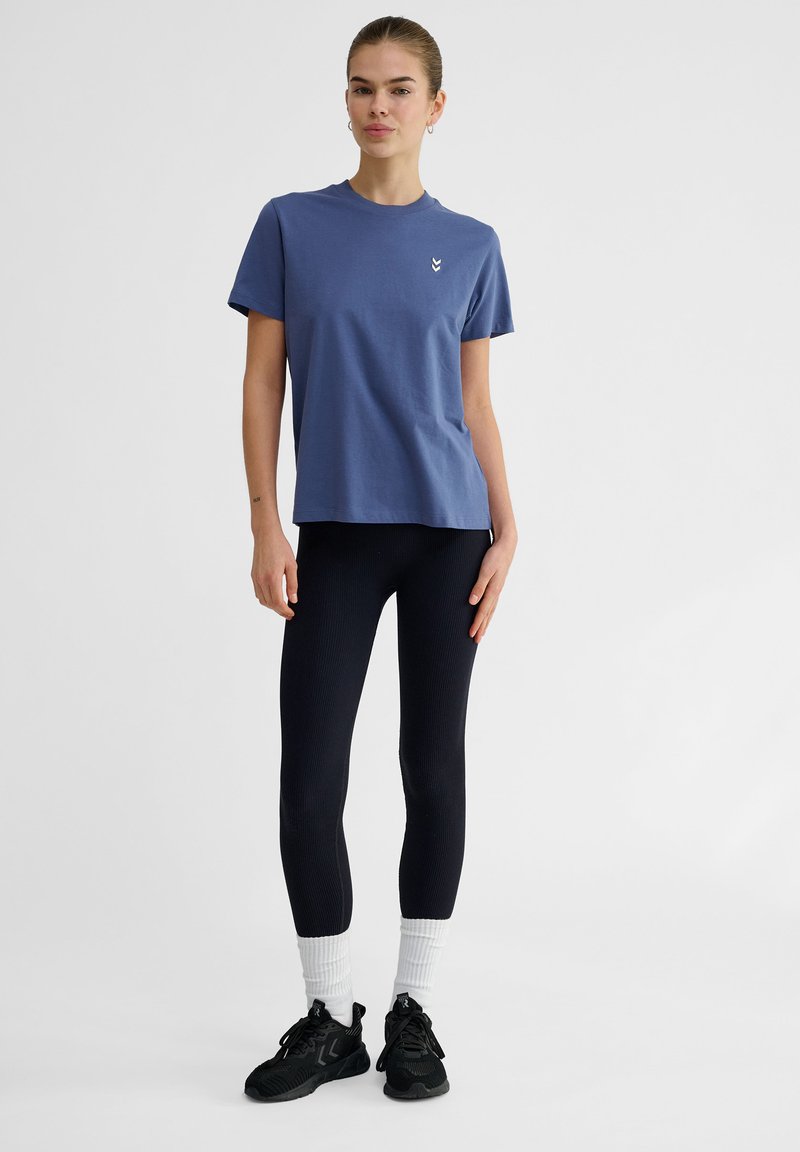 Blå bomullströja med korta ärmar, prydd med en liten vit logotyp, i kombination med svarta ribbade leggings och svarta träningsskor.