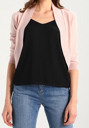 Mujer con cárdigan corto rosa claro sobre top negro sin mangas y vaqueros azules, mostrada desde los hombros hasta las caderas, de pie sobre fondo blanco.