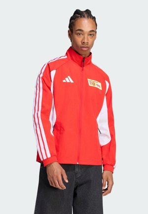 Jeune homme portant une veste de sport zippée rouge et blanche avec des manches rayées et des logos, associée à un pantalon noir.