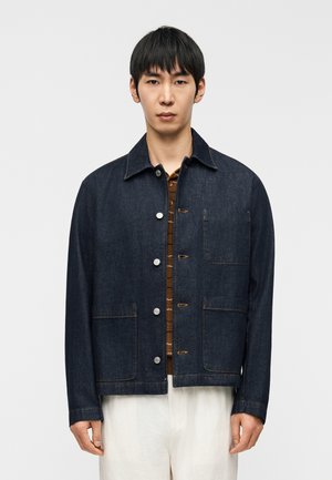 Tamsiai mėlynos spalvos džinsinė striukė su apykakle, didelėmis kišenėmis su atvartais ir sidabriniais mygtukais. Turi kvadratinį kirpimą ir kontrastingas siuvimo detales.