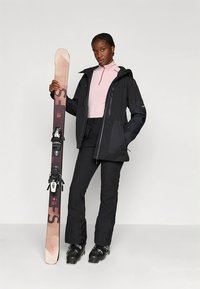 Giacca da sci nera e pantaloni con cerniere, indossati sopra una maglia a maniche lunghe rosa chiaro; tiene uno sci con attacchi; texture lisce.