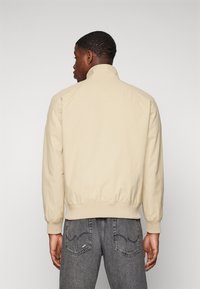 Chaqueta bomber beige con un alto cuello, puños acanalados y un dobladillo ajustado. El material parece suave. Llevada con jeans gris oscuro que presentan acentos de costura en los bolsillos.