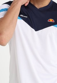 Muž v bílém a námořnickém atletickém tričku s modrými akcenty, upravující si límec; tričko má na hrudi loga značky ellesse.