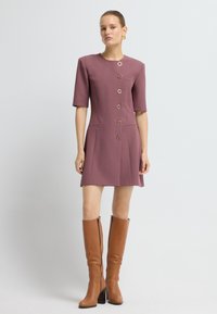 Vestido de línea A de mangas cortas en rosa empolvado con escote redondo, que presenta botones decorativos y bolsillos laterales. Combinado con botas marrones hasta la rodilla.