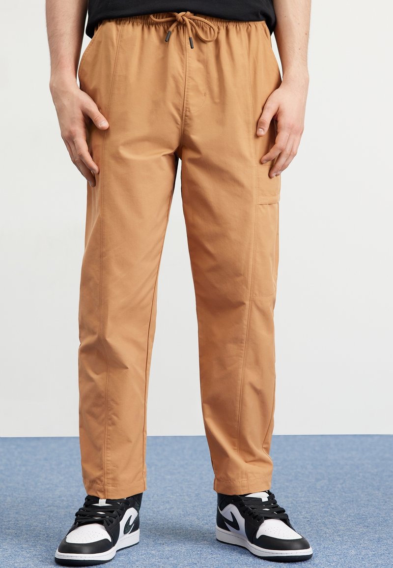 Personne portant un pantalon beige à cordon et des baskets noir et blanc, debout sur un tapis bleu, les mains détendues le long du corps.