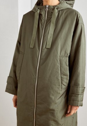 Personne portant une parka à capuche vert olive avec fermeture éclair à l'avant, grandes poches et cordons pendants de la capuche.