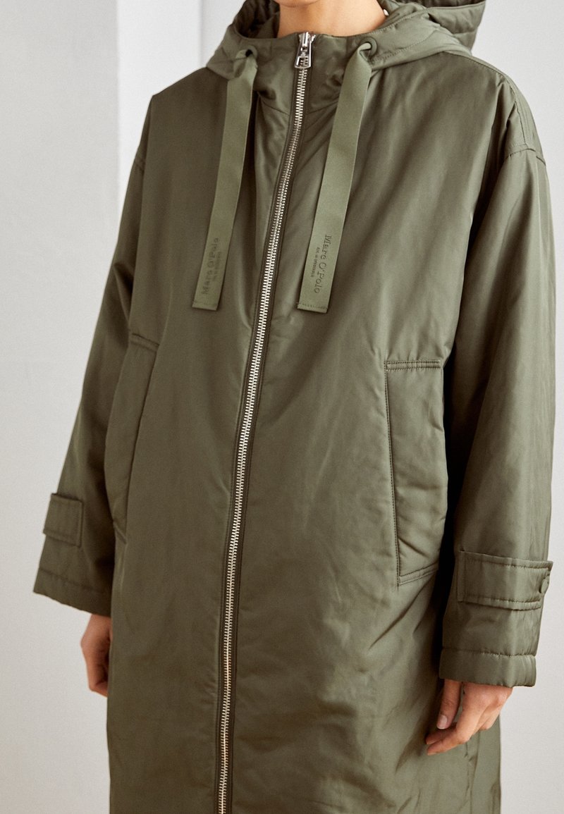 Personne portant une parka à capuche vert olive avec fermeture éclair à l'avant, grandes poches et cordons pendants de la capuche.