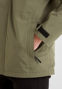 Olive green jacket with a smooth texture, featuring a side pocket, Velcro cuff closure, and a straight hem. Minimalistic design.