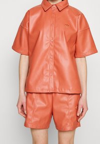 Camicia in ecopelle corallo a maniche corte con colletto e bottoni a pressione, abbinata a pantaloncini coordinati con bottoni laterali e dettagli con logo.