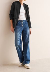 Veste bomber noire avec des poignets côtelés, T-shirt blanc, jeans en denim bleu taille haute avec fermeture à boutons, et baskets blanches.