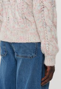 Pulover tricotat în nuanțe pastelate, cu un model de cablu și margine rib, asociat cu blugi din denim albastru, evidențiind textura și contrastul de culoare.