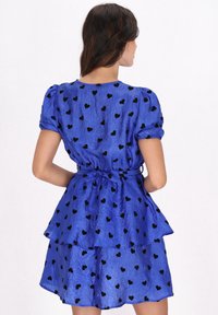 Blaues Kleid mit schwarzen Herzmustern, gestuftem Design, elastischen Puffärmeln und einem Bindegürtel in der Taille. Glatter, leicht glänzender Stoff.