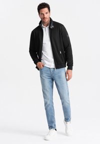 Giacca nera con zip e finitura texturizzata, polo bianca sotto, jeans azzurri chiari e sneakers bianche con accenti neri.