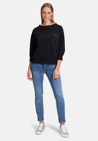 Sweat-shirt noir avec un col bateau, présentant des lettres et des motifs texturés. Associé à un jean bleu clair et des baskets métalliques.