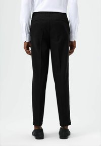 Zwarte, op maat gemaakte broek met een gladde textuur, elastische tailleband en twee achterzakken. Gecombineerd met een wit shirt met lange mouwen.