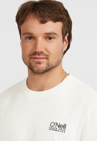 Bílá bavlněná tričko s kulatým výstřihem a vyšitým černým logem na levém prsu, které zní "O'Neill Santa Cruz California U.S.A."
