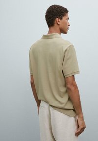 Beige polo tričko s uvolněným střihem, krátkým rukávem a měkkou texturou. Obsahuje klasický límec a jemné švy.