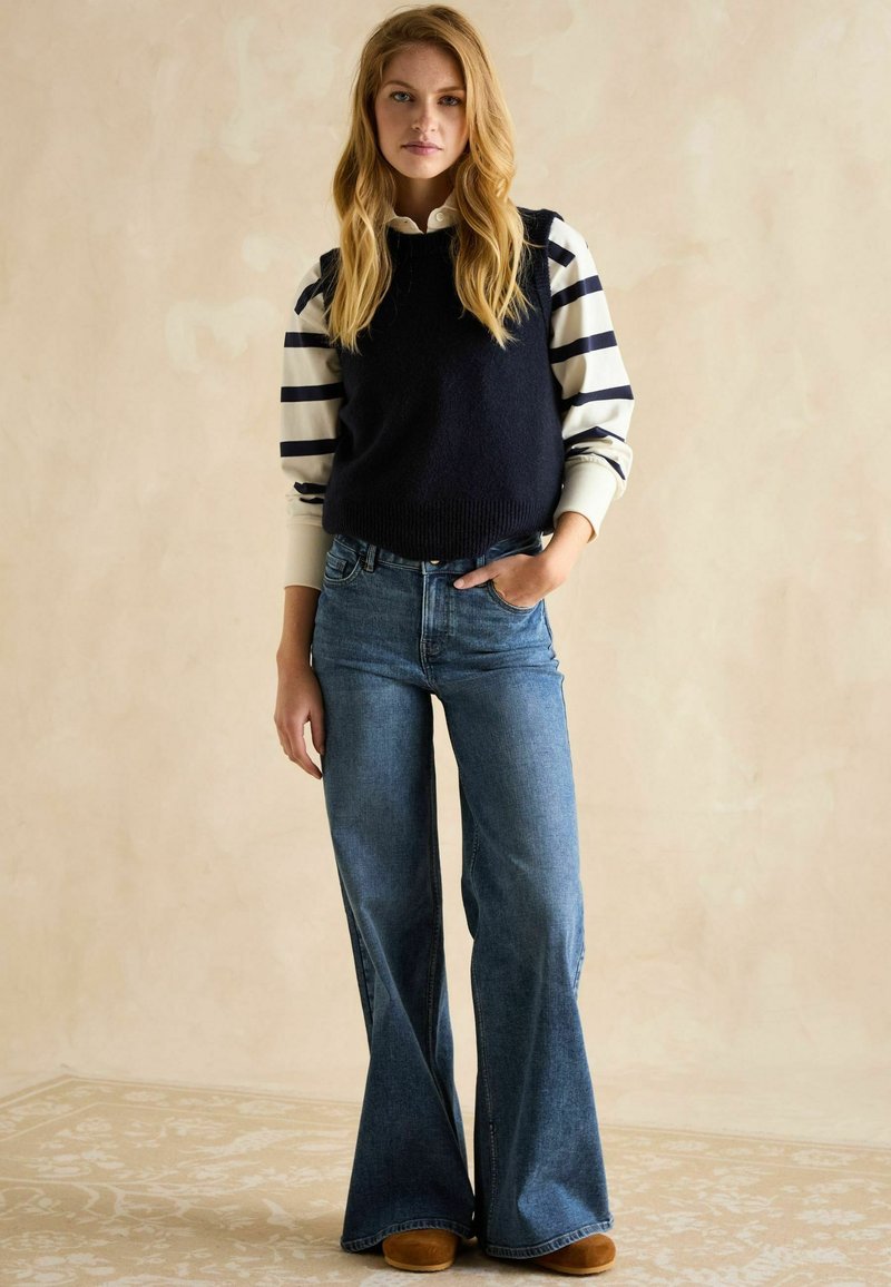 Marineblauer Strick-Weste über einem weiß und marineblau gestreiften Langarmhemd, kombiniert mit weit geschnittenen blauen Jeans und beigen Schuhen. Neutrale Hintergrund.