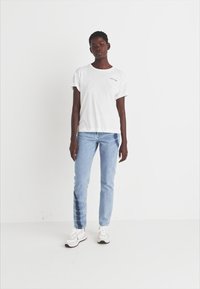 T-shirt branca de algodão com mangas curtas e decote redondo. Jeans de ganga azul com um lavagem clara e um detalhe único de tie-dye numa perna. Ténis brancos.