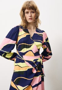 Wickelkleid mit abstrakten Mustern in Marineblau, Pink und Grün. Weicher Stoff, gepuffte Ärmel und ein seitliches Bindemdetail betonen die Form.